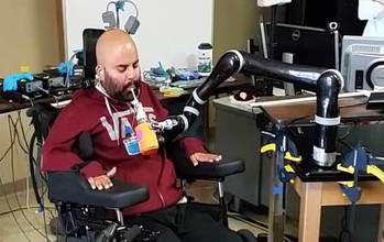 Man in a chair drinking from a cup held by a robotic arm